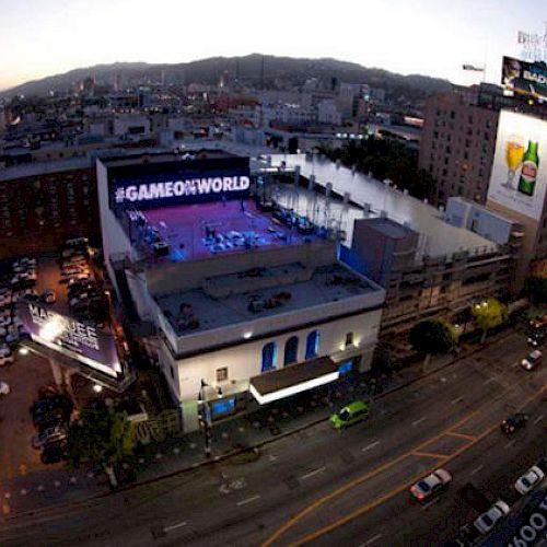 The image shows a cityscape during twilight with streets, buildings, and a large advertisement that reads "#GAMEONWORLD" on one of the rooftops.