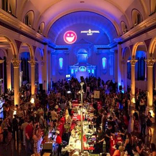 A large indoor event in an ornate hall with arched columns, featuring numerous people socializing, food or drink stations, and ambient lighting.