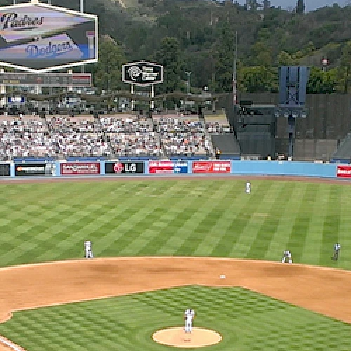 A baseball stadium with a game in progress, featuring green grass, advertisements, and a crowd of spectators in the stands.
