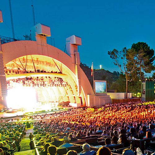 An open-air amphitheater filled with an audience enjoying a vibrant, illuminated stage show during the evening.