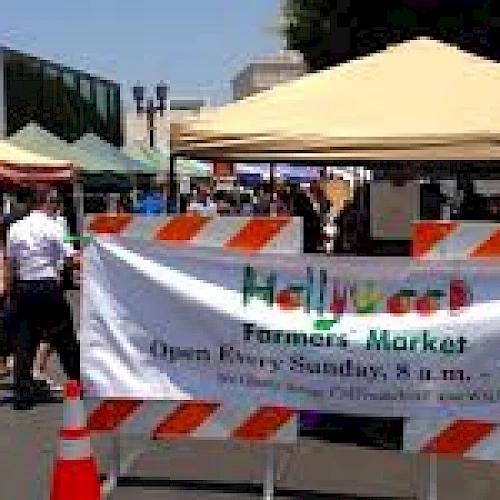 A farmers market with multiple stalls under tents, and a sign reading "Hollywood Farmers Market, Open Every Sunday, 8 a.m. - 1 p.m." Ends the sentence.