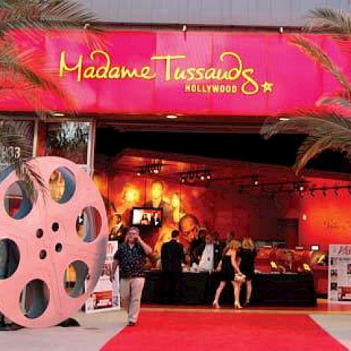 The image shows the entrance to Madame Tussauds Hollywood with a red carpet, palm trees, large film reels, and people entering the building.