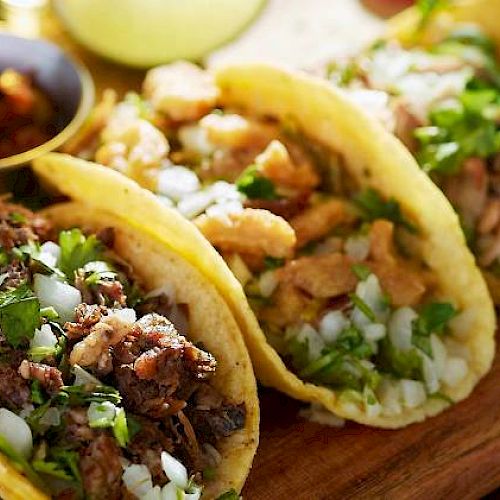 The image shows three tacos filled with various meats, topped with chopped onions and cilantro, served on a wooden board with a side of salsa.