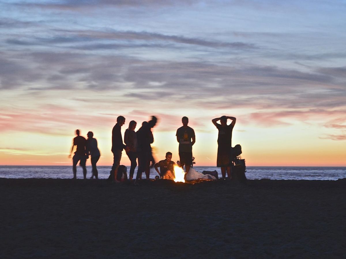 A group of people stands around a campfire on a beach during sunset with the ocean in the background.