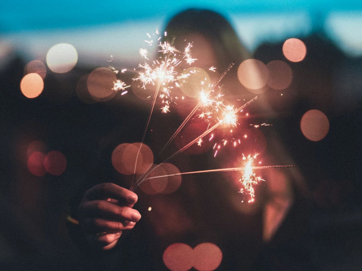 A person holding multiple sparklers with bright, glowing lights in the background, creating a festive atmosphere in a blurred night scene.