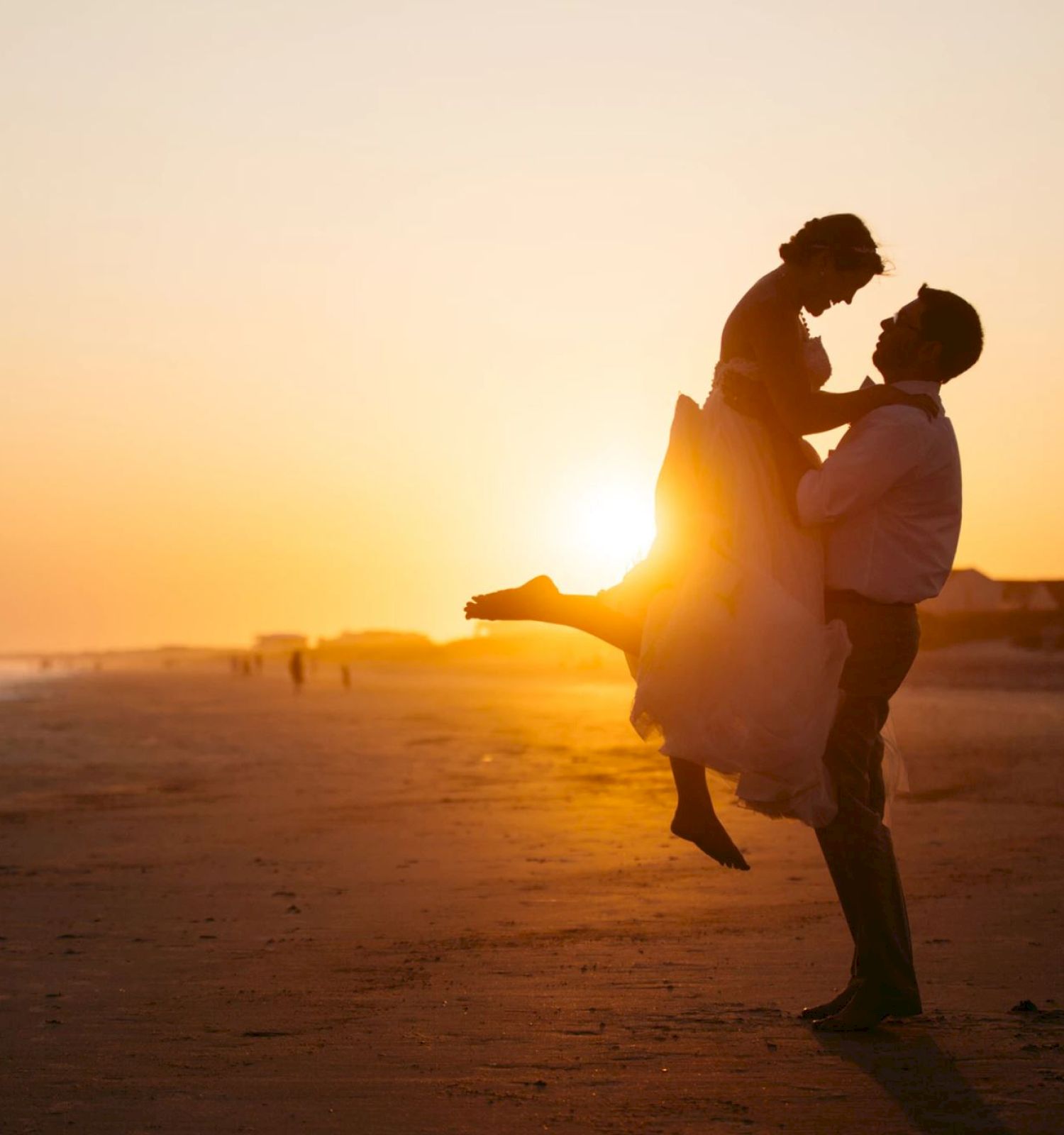 A couple is on a beach at sunset, with the man lifting the woman in a joyful pose. Houses and distant figures are visible in the background.