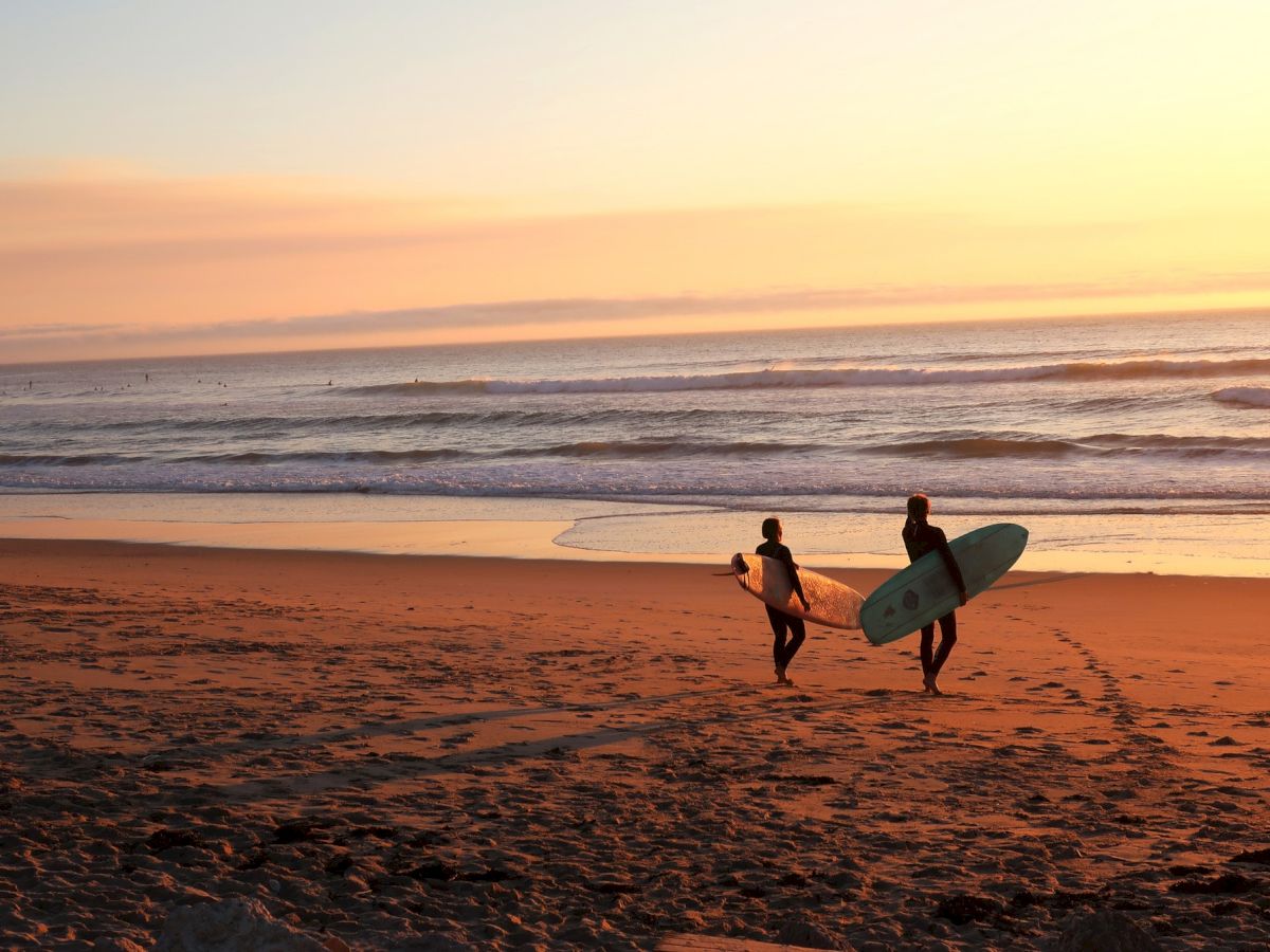 Two surfers walk along the beach at sunset, carrying their surfboards towards the ocean as waves roll in the background.