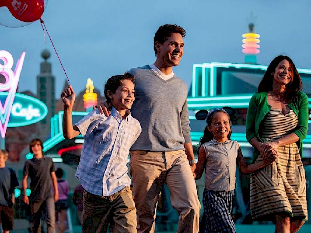 A family of four is joyfully walking, with neon lights in the background, and the boy is holding a red balloon.