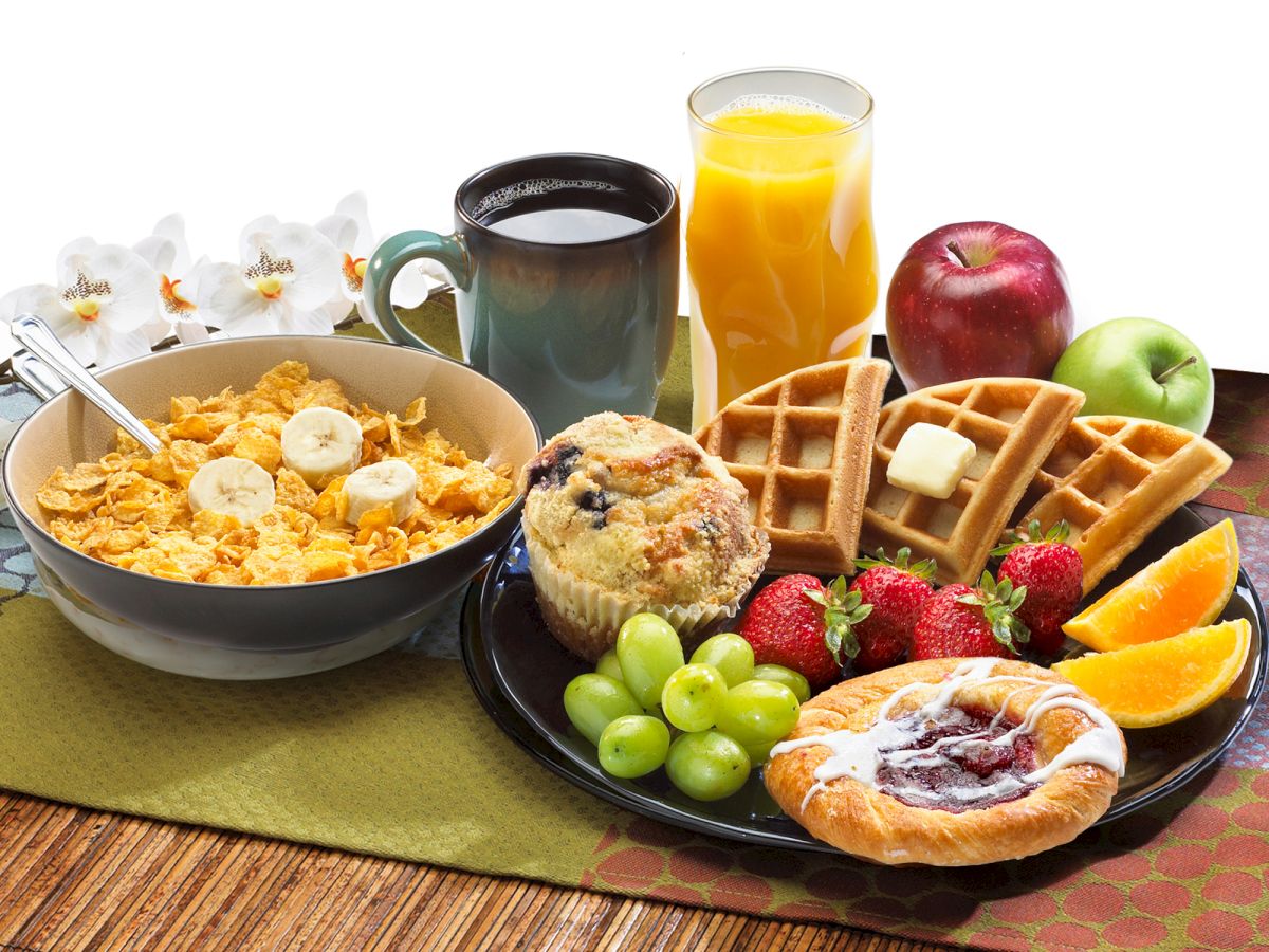 The image shows a breakfast spread with cereal, bananas, waffles, muffin, fruit, coffee, and orange juice on a table setting.