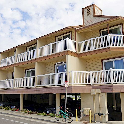 A three-story building with multiple balconies, white railings, and ground-level parking. A bicycle is parked near the building under a blue sky.