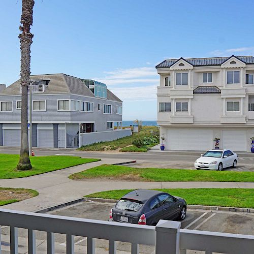 A view from a balcony showing a parking area with cars, a grassy space, palm trees, and residential buildings near the coast under a clear sky.