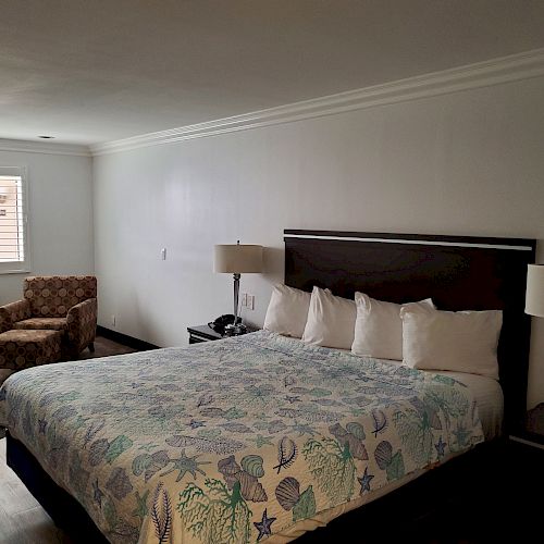 The image shows a tidy hotel room with a neatly made king-size bed, two bedside tables with lamps, a patterned armchair, and a window.