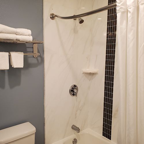 A modern bathroom with a wall-mounted shelf holding towels and a tub-shower combination with a white curtain and marble tile accents.