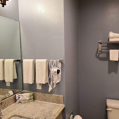 A small bathroom with a mirror, towels, a granite countertop, a sink, a hairdryer, a toilet, and towel racks on a light blue wall.