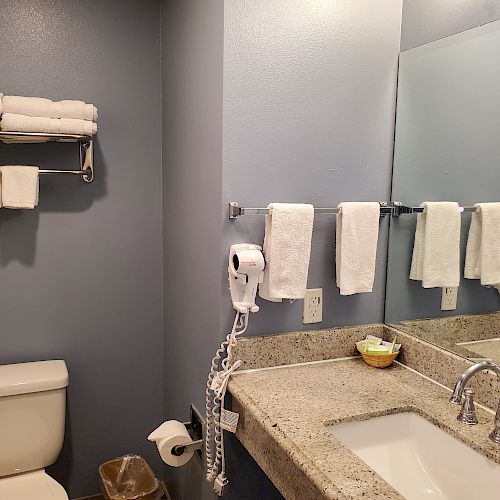 The image shows a bathroom with towels, a sink, a mirror, a toilet, a hairdryer mounted on the wall, and a waste bin.