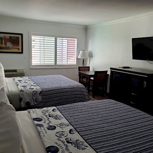 A hotel room with two neatly made beds, a wall-mounted TV, a desk with a lamp, a framed artwork, and a window with blinds is shown.