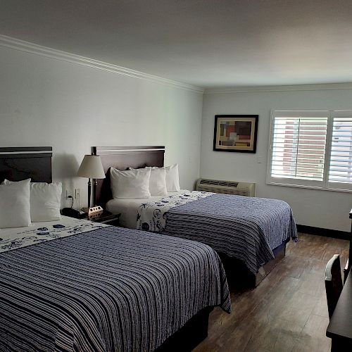 A hotel room with two double beds, striped bedspreads, a desk, lamps, a window, and a framed picture on the wall.