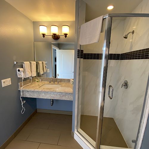 A bathroom with a glass shower, dual sinks, wall-mounted hair dryer, towels, and stylish lighting above the mirror, giving it a modern look.