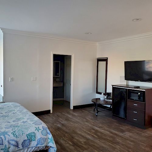 A hotel room with a bed, wall-mounted TV, mini-fridge, mirror, small table, and bathroom entrance, decorated in a nautical theme.