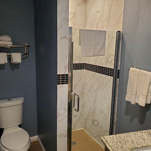 The image shows a bathroom with a shower, toilet, towel rack with towels, wall-mounted hairdryer, and sink with countertop and toiletries.