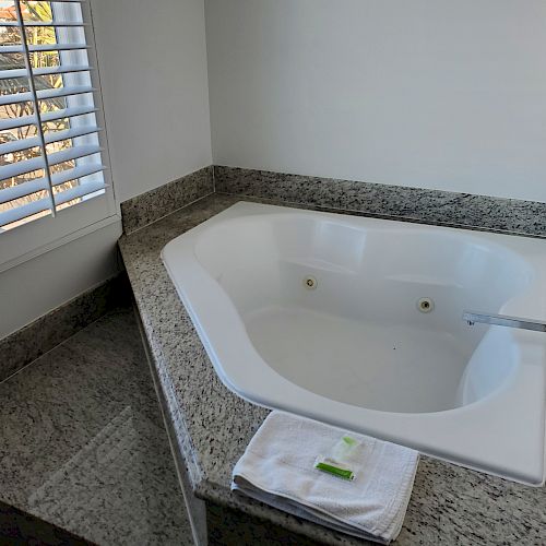 A corner bathtub with granite surrounds, an open window with shutters, and a folded towel with a small soap bar on the counter are shown.