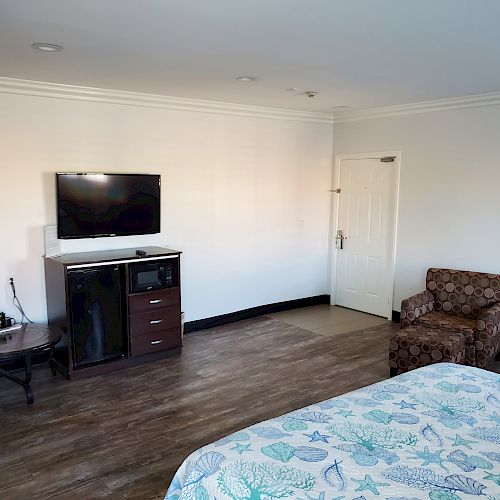 The image shows a hotel room with a bed, a TV, a microwave, a small refrigerator, a table, a chair, and a door. The floor appears to be wood.