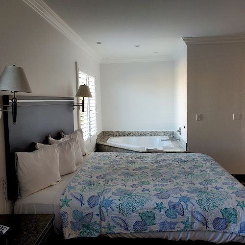 This image features a hotel room with a bed, pillows, bedspread with sea patterns, bedside lamps, a window with shutters, and a hot tub in the corner.