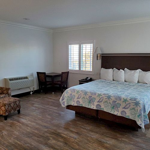 A spacious room with a large bed, floral bedspread, two brown chairs, a small table with chairs, a window, and wood flooring.