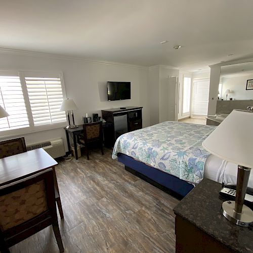 A cozy hotel room featuring a bed with floral bedding, a desk, chairs, lamps, a dresser with a TV, and large windows with blinds.