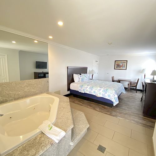 The image shows a hotel room featuring a bed, a jacuzzi tub, a desk, a flatscreen TV, and a dresser with a mirror in a bright, clean setting.