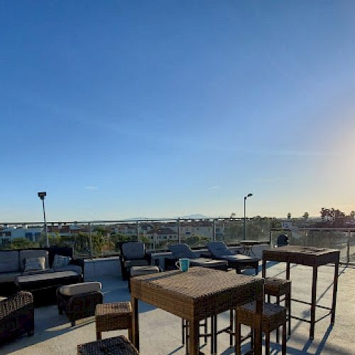 The image shows a rooftop patio with wicker furniture, tables, and chairs under a clear blue sky and the sun shining in the background.