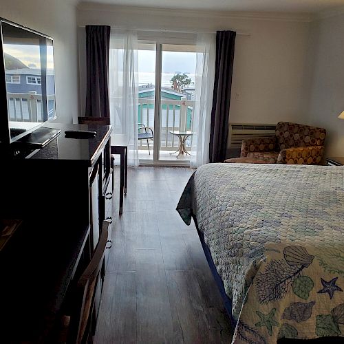 A cozy hotel room features a bed with a patterned quilt, a TV, a small armchair, a lamp, and a balcony with chairs and an ocean view.