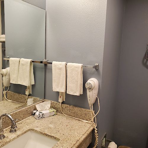 A bathroom with a granite countertop, sink, mirror, hair dryer, towels on racks, marble shower, toilet, and tissue box.