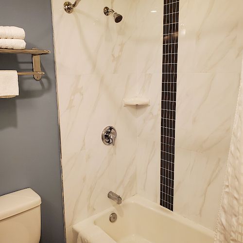 A bathroom with a bathtub and shower, white marble tiles, a toilet, and a towel rack holding folded towels and washcloths on a blue-gray wall.