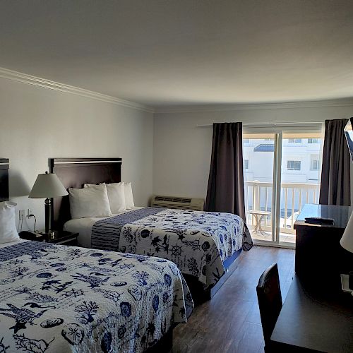 A hotel room with two queen beds, sea-themed bedding, a TV, a desk, a lamp, and a sliding door leading to a balcony or patio area.