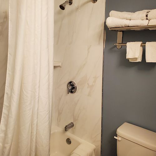 A bathroom with a marble-tiled shower, white shower curtain, toilet, and wall-mounted shelf holding folded towels and washcloths against a blue wall.