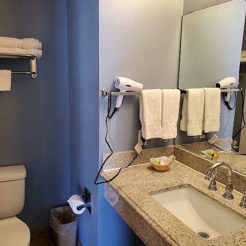 The image shows a bathroom with a toilet, mounted towel racks, a sink with a granite countertop, a mirror, wall-mounted hairdryer, and various toiletries.