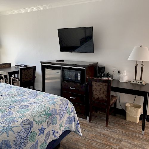 The image shows a hotel room with a bed, TV, mini fridge, microwave, two chairs, a desk, and table lamps on dark wood furniture. The room has a cozy feel.