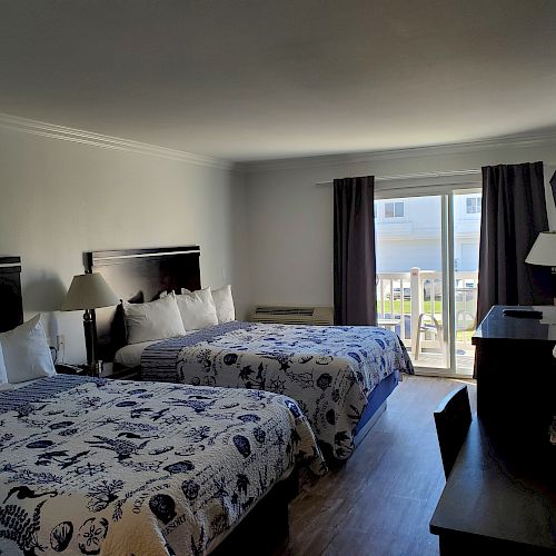 A well-lit hotel room with two double beds features ocean-themed bedding, a work desk, a TV, dark curtains, and a sliding door leading to a balcony.