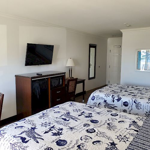 A hotel room with two beds, a wall-mounted TV, a dresser, a desk with a chair, a lamp, and a mirror next to the door and window.