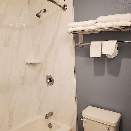 A bathroom with a bathtub, marble wall, and fixtures, alongside towels on a metal rack above a toilet against a gray wall.