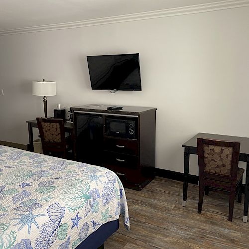 The image shows a hotel room with a bed, a TV mounted on the wall, a dresser with a microwave and fridge, a lamp, and a small table with two chairs.