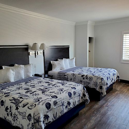 The image shows a hotel room with two double beds, each with patterned bedding, a wooden floor, a window with blinds, and a wall-mounted lamp.