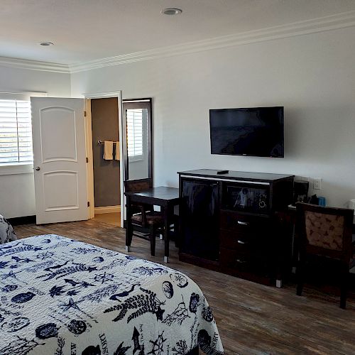 A hotel room with two beds, a wall-mounted TV, desk, chair, large window with blinds, and a bathroom door to the left are visible.