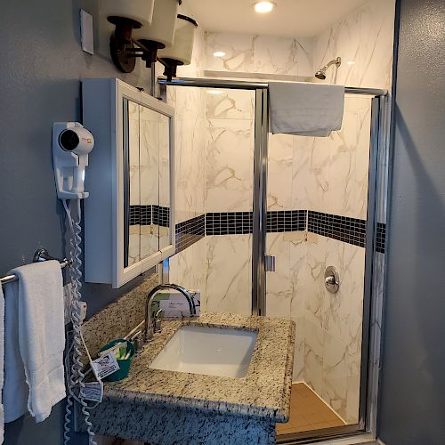 The image shows a bathroom with a granite countertop sink, mirror, wall-mounted hair dryer, towel rack, and a shower with a glass door.