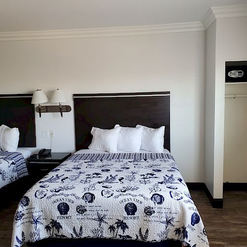 A hotel room with two beds featuring patterned quilts, a nightstand with a lamp, and a closet space with a safe and a hanger.