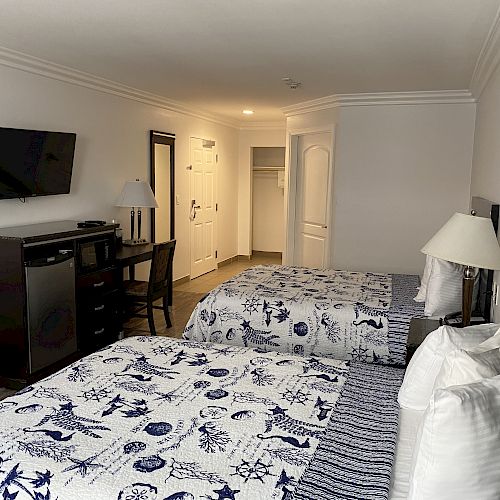 The image shows a hotel room with two beds, a TV, a dresser, a desk with a chair, lamps, and a white-themed decor.