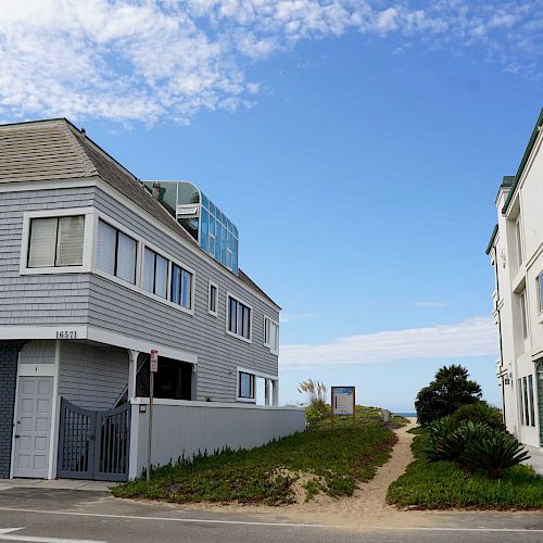 The image shows two residential buildings flanking a sandy path that leads to the beach, set under a sky with some scattered clouds.