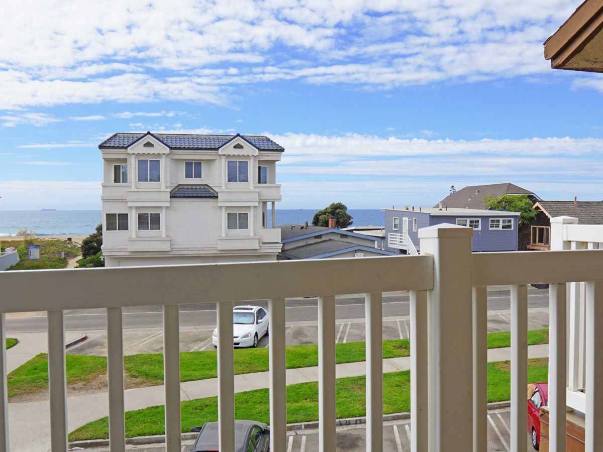 A view from a balcony, showing a residential area with houses, green lawns, a parked car, and the ocean in the background under a partly cloudy sky.