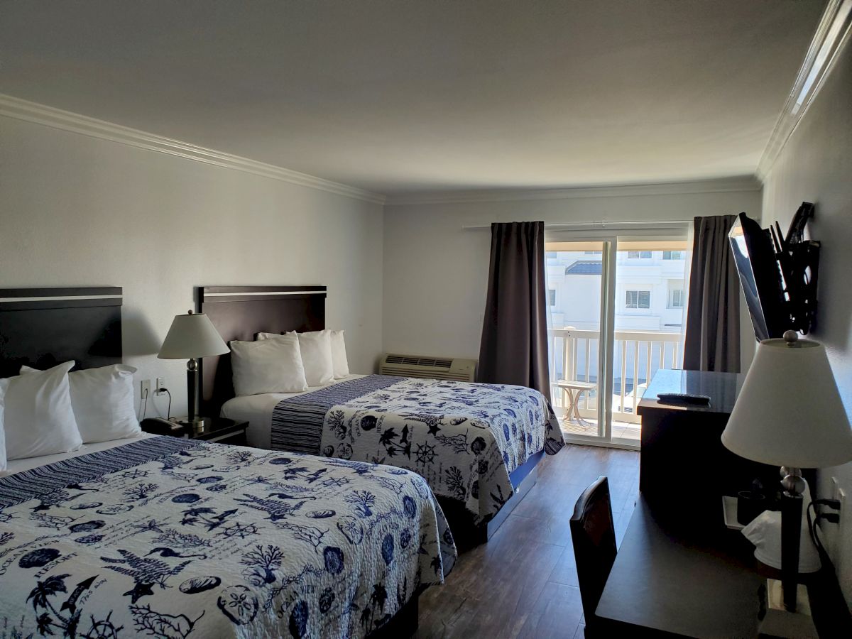 A hotel room with two double beds, a desk, a wall-mounted TV, a lamp, and a balcony with a view outside.